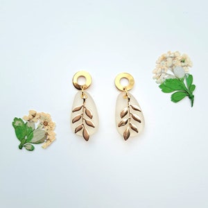 May include: Gold-coloured earrings with a leaf design. Each earring has a gold circle at the top, a frosted teardrop-shaped element, and a gold leaf charm. The earrings are displayed on a white background with dried flowers.