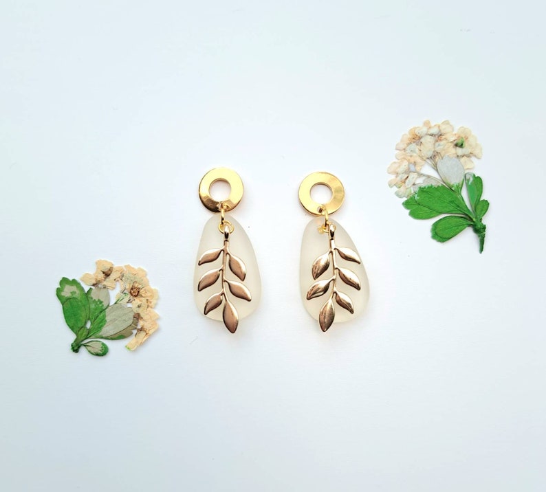 May include: Gold-coloured earrings with a leaf design. Each earring has a gold circle at the top, a frosted teardrop-shaped element, and a gold leaf charm. The earrings are displayed on a white background with dried flowers.