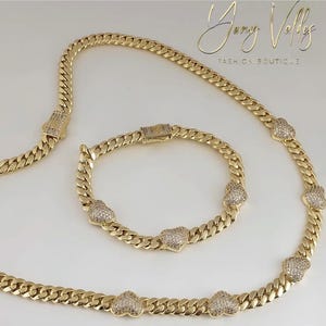 Op de afbeelding: Een goudkleurige sieradenset met een ketting en armband. De ketting en armband hebben een gourmette kettingontwerp met hartvormige accenten bezet met kleine, sprankelende stenen. De set wordt gepresenteerd op een wit oppervlak.