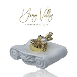 Puede incluir: Un anillo dorado con forma de abeja con cristales transparentes y una perla en el centro, colocado sobre un pedestal decorativo blanco. El anillo presenta diseños detallados. El texto "Jany Valley Fashion Boutique" está arriba.