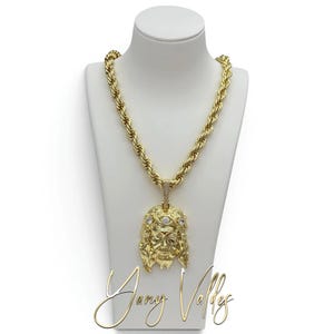 Puede incluir: Un collar de cadena de cuerda en tono dorado con un gran colgante en forma de rostro con una corona de espinas. El colgante está adornado con pequeñas piedras transparentes incrustadas en la corona y el asa, con una textura metálica detallada.