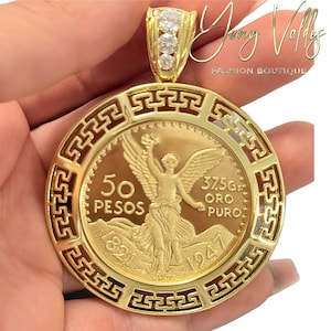 Puede incluir: Un colgante de oro con una moneda de 50 Pesos Oro Puro de 1947, enmarcada en un diseño circular con patrón de greca. El pasador está adornado con tres piedras claras. La moneda muestra una figura alada y el texto 50 PESOS 37.5Gr.