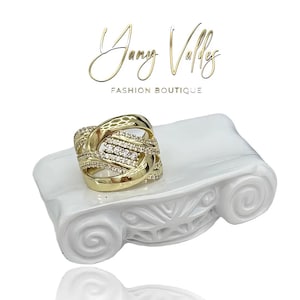 Puede incluir: Un anillo dorado con un diseño entrelazado, adornado con pequeñas piedras preciosas transparentes. El anillo se exhibe sobre un pedestal blanco decorativo. El texto "Jany Vallys Fashion Boutique" está en letras doradas arriba.