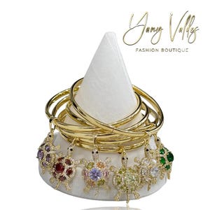 Puede incluir: Brazaletes dorados con colgantes de tortuga adornados con piedras preciosas de colores. Los brazaletes se exhiben en un soporte blanco en forma de cono. El texto "Yany Valley Fashion Boutique" es visible.
