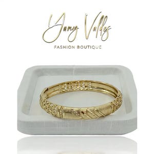 Puede incluir: Un brazalete dorado con intrincados diseños grabados. El brazalete se exhibe en una bandeja blanca. El texto "Jany Valley Fashion Boutique" está encima del brazalete.