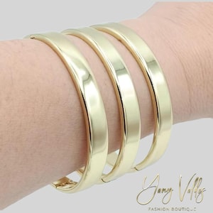 Puede incluir: Cuatro brazaletes dorados de banda plana apilados en una muñeca. Los brazaletes tienen un acabado liso y pulido y un diseño simple y elegante. Las joyas se exhiben sobre un fondo gris claro.
