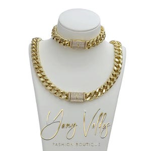 Könnte beinhalten: Ein goldfarbenes Kettenhalsketten- und Armband-Set. Beide Teile weisen ein dickes Panzerketten-Design mit einem rechteckigen Verschluss auf, der mit kleinen, klaren Steinen verziert ist. Der Schmuck wird auf einer weißen Schaufensterpuppe präsentiert. Der Text "Jany Vollez Fashion Boutique" steht unten.