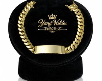 Puede incluir: Una pulsera de cadena de eslabones cubanos en tono dorado con una placa de identificación rectangular y lisa en el centro. La pulsera tiene un cierre seguro y se muestra sobre un soporte de terciopelo negro con el texto Yany Valdes Fashion Boutique Premium.