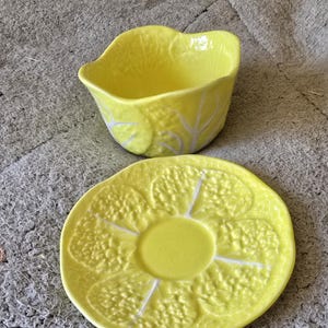 May include: A vibrant yellow ceramic bowl and saucer. The bowl is shaped like a leaf with white vein detailing. The round saucer features a textured cabbage leaf design. The set is displayed on a grey surface.