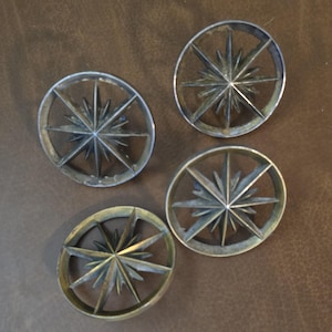 May include: Four circular metal objects with a starburst design. The objects have a wheel-like appearance with spokes radiating from a central star. The metal has a silver and brass finish, and they are arranged on a brown surface.