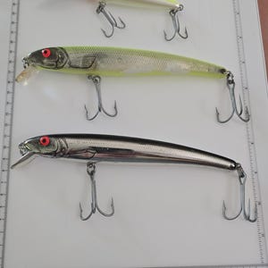 May include: Three fishing lures placed on a white cutting mat with a ruler. The top lure is yellow and white, the middle is green and silver with red eyes, and the bottom is metallic silver with red eyes. Each lure features two metal treble hooks.