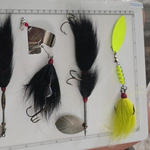 May include: Four fishing lures arranged on a white measuring board. Three lures feature black fur tails, silver or metallic blades, and treble hooks. The fourth lure has a bright neon yellow blade, yellow beads, and a neon yellow fur tail.