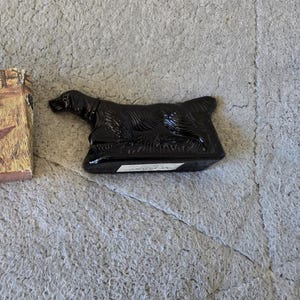 Può includere: Una bottiglia nera a forma di cane accanto a una scatola con una scena di caccia. La scatola presenta illustrazioni di cani, uccelli e cacciatori in un campo. La bottiglia è lucida e probabilmente in vetro o ceramica.