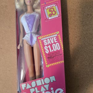 May include: A Barbie doll in a pink and white box. The doll has blonde hair and is wearing a white and purple outfit. The box has text that says "Fashion Play Barbie" and "Save $1.00".