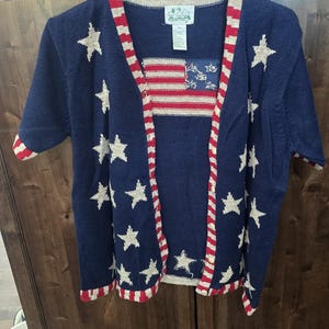 May include: A navy blue cardigan sweater with white stars and a red, white, and blue American flag design. The sweater has short sleeves and a striped trim. The sweater is hanging on a white hanger.