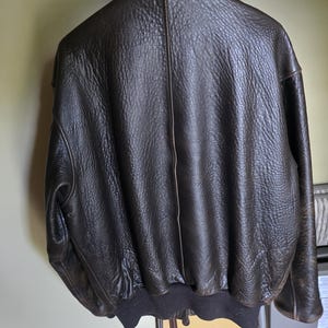 May include: A dark brown leather bomber jacket hanging on a white wire rack. The jacket has a textured surface, a ribbed hem, and a classic collar. The back of the jacket is visible, showcasing its design and stitching.