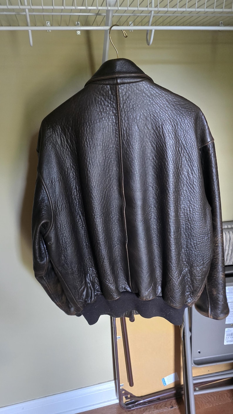 May include: A dark brown leather bomber jacket hanging on a white wire rack. The jacket has a textured surface, a ribbed hem, and a classic collar. The back of the jacket is visible, showcasing its design and stitching.