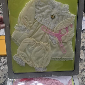 May include: A vintage baby doll outfit set, featuring a cream-colored dress and bloomers with lace trim, packaged in a Dreamland Series box. The outfit is designed to fit 16-18 inch dolls. Includes a pink hanger.