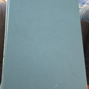 May include: A closed, light blue book with a slightly worn cover. The book is rectangular and appears to be hardcover. The edges show minor wear and tear. The book is resting on a soft, pink fabric.
