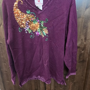 Pode incluir: Um suéter cor de ameixa com decote em V e mangas compridas. O suéter apresenta um design de lantejoulas em tons de dourado, laranja, verde e roxo na frente. A etiqueta da marca diz "Quacker Factory". A bainha e os punhos têm uma borda com franjas.