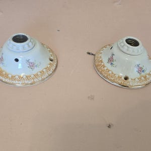 May include: Two vintage white ceramic ceiling light fixtures with a floral design and gold trim. Each fixture has a threaded socket and mounting holes. The fixtures are on a light brown surface.