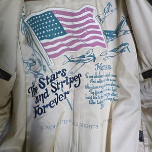 May include: Brown leather jacket with a cream-colored lining. The lining features an American flag and text that reads "The Stars and Stripes Forever." The jacket has a classic design with a label that reads "TYPE A-2".