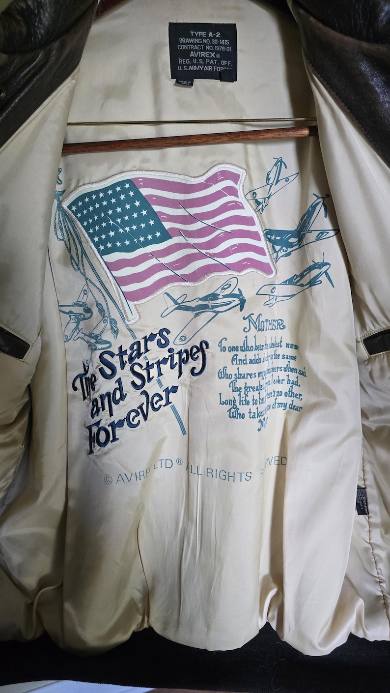 May include: Brown leather jacket with a cream-colored lining. The lining features an American flag and text that reads "The Stars and Stripes Forever." The jacket has a classic design with a label that reads "TYPE A-2".