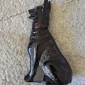 May include: A glossy, dark purple figurine of a German Shepherd dog in a seated position. The dog has pointed ears and a collar. The figurine is made of a shiny material and sits on a textured, gray surface.