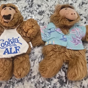 May include: Two brown plush ALF dolls. One doll wears a white apron with blue text that reads "Cookin' with ALF". The other doll wears a blue shirt with a floral pattern. Both dolls have a tan snout and brown fur.