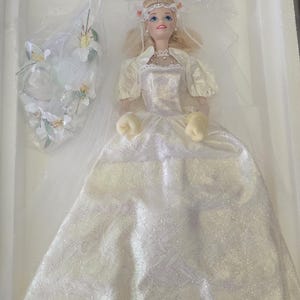 Peut inclure: Poupée blonde vêtue d'une robe de mariée blanche avec un voile, présentée dans une boîte blanche. La robe présente un motif scintillant et des manches bouffantes. Un bouquet de fleurs blanches est dans un sac en plastique. La poupée porte un diadème.
