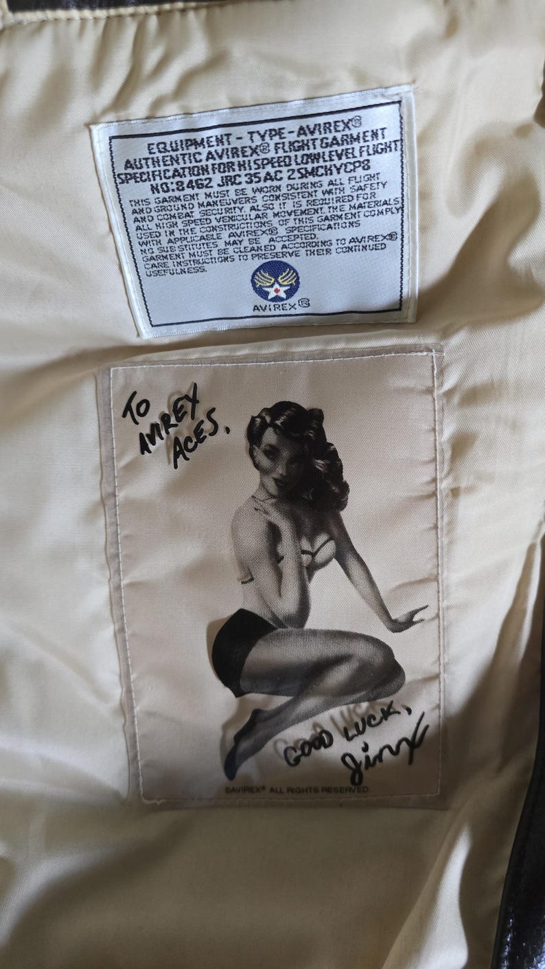 May include: Close-up of the interior of a vintage Avirex flight jacket. The lining features a printed pin-up girl graphic with the text "Good Luck, Jinx" and "To Avirex Aces." A white label displays technical specifications.