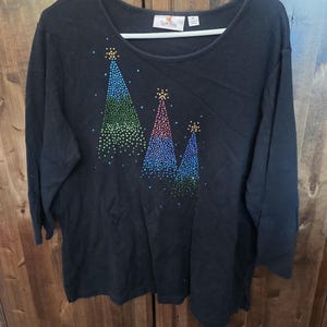 以下が含まれることがあります： 3つのクリスマスツリーのデザインが施された黒い長袖シャツ。各ツリーはカラフルでキラキラ光るドットで飾られています。ツリーは青、緑、赤の色合いで、上部に金色の星のアクセントが付いています。