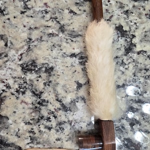 May include: A handcrafted wooden pipe with a light-coloured fur wrap and decorative bands of red, orange, and yellow. A leather cord is attached to the base. The pipe is set against a speckled background.