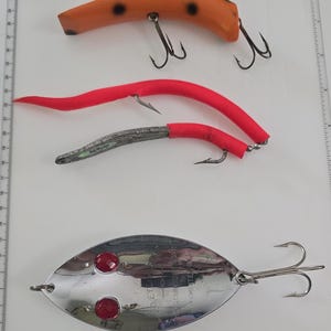 May include: Four fishing lures arranged on a white surface with a ruler border. One orange lure with black spots, two red soft plastic lures with hooks, and one large, oval, silver-colored metal spoon lure with two red eyes and text.