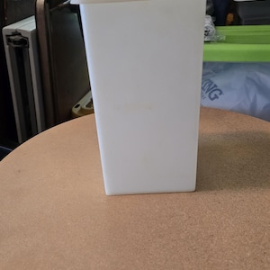 Puede incluir: Un recipiente rectangular blanco con una tapa a juego. El recipiente está sobre una mesa redonda y marrón. El fondo incluye una calabaza dorada decorativa y otros objetos.