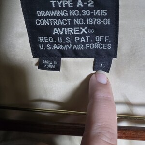 May include: Close-up of a black label with white text from an A-2 jacket. The label reads "TYPE A-2", "DRAWING NO. 30-1415", "CONTRACT NO. 1978-01", and "AVIREX&reg;". It also states "REG. U.S. PAT. OFF. U.S. ARMY AIR FORCES" and "MADE IN KOREA". A finger points to the "L" size tag.