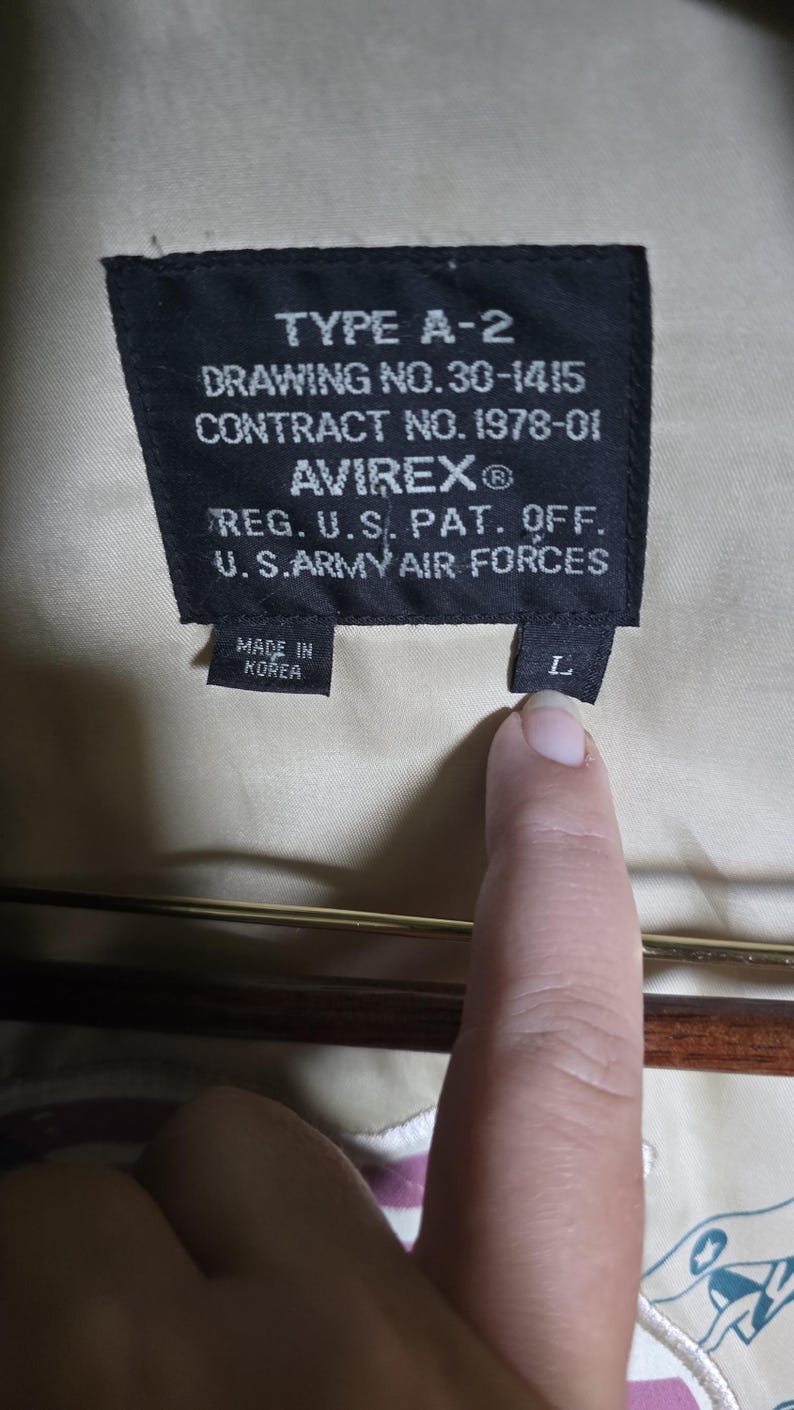 May include: Close-up of a black label with white text from an A-2 jacket. The label reads "TYPE A-2", "DRAWING NO. 30-1415", "CONTRACT NO. 1978-01", and "AVIREX&reg;". It also states "REG. U.S. PAT. OFF. U.S. ARMY AIR FORCES" and "MADE IN KOREA". A finger points to the "L" size tag.