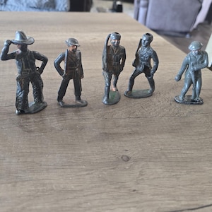 Puede incluir: Un conjunto de seis pequeñas figuras de juguete de metal antiguas que representan varias figuras. Las figuras están pintadas en tonos grises y negros, y están posando en diferentes posturas. Las figuras están dispuestas sobre una superficie de madera.