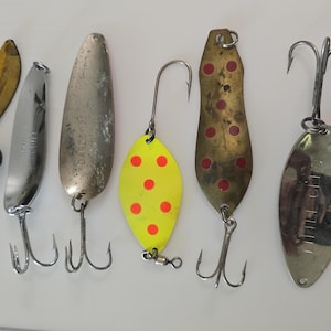 May include: A collection of seven metal fishing lures arranged vertically. The set includes various spoon-shaped designs in gold, silver, and yellow with red dots, featuring treble hooks, a black weight, and a small detached amber-colored leaf piece.