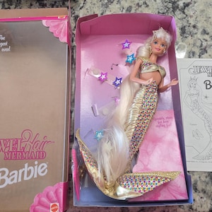 Puede incluir: Una muñeca Barbie Sirena de pelo joya en su caja original. La muñeca tiene pelo largo rubio, una cola de sirena dorada y con lentejuelas, y un top dorado. La caja es rosa y tiene el logo de Barbie y las palabras "Jewel Hair Mermaid".