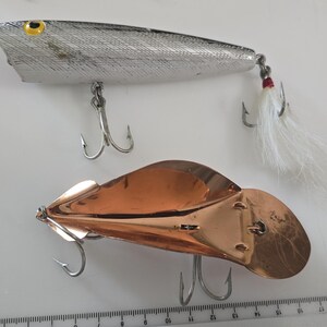 May include: Two fishing lures positioned next to a ruler showing measurements in inches. The top lure is silver with a yellow eye and white feather tail. The bottom lure is a polished copper spoon-style lure with two metal treble hooks.
