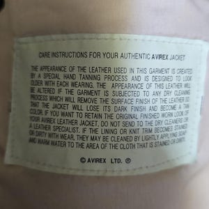 May include: Close-up of a cream-colored leather jacket interior with a care instructions label. The label provides instructions for cleaning and maintaining the jacket's appearance. The jacket has a serial number and the Avirex Ltd. logo.