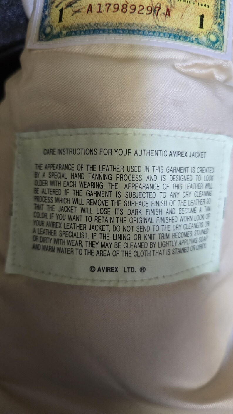 May include: Close-up of a cream-colored leather jacket interior with a care instructions label. The label provides instructions for cleaning and maintaining the jacket's appearance. The jacket has a serial number and the Avirex Ltd. logo.