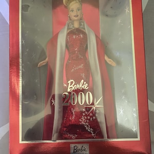 May include: A Barbie doll from the year 2000, in a red sequined gown and white shawl, displayed in a red collector's edition box. The box has the Barbie logo and the text "Barbie 2000".