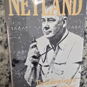 May include: A book cover featuring a black and white illustration of a man holding a whistle. The title "NEYLAND" is in gold letters. The book is titled "The Gridiron General" by Bob Gilbert. The background has football play diagrams.