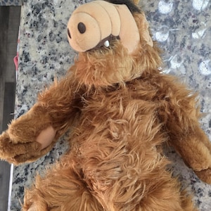 Puede incluir: Un peluche del personaje Alf, con un hocico marrón, ojos marrones y pelaje marrón hirsuto. El juguete tiene una barriga y brazos de color marrón claro, y está tumbado sobre una encimera moteada.
