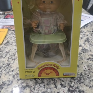 May include: A boxed "Walking Doll" toy from Sunshine Kids. The doll is wearing a pink dress and sitting in a green high chair. The box is yellow with red hearts and the text "Walking Doll".