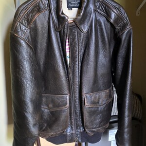 May include: A brown leather jacket with a zip-up front, two front pockets, and a ribbed hem and cuffs. The jacket is hanging on a hanger in a closet. The jacket has a collar and a label that reads "TYPE A-2".