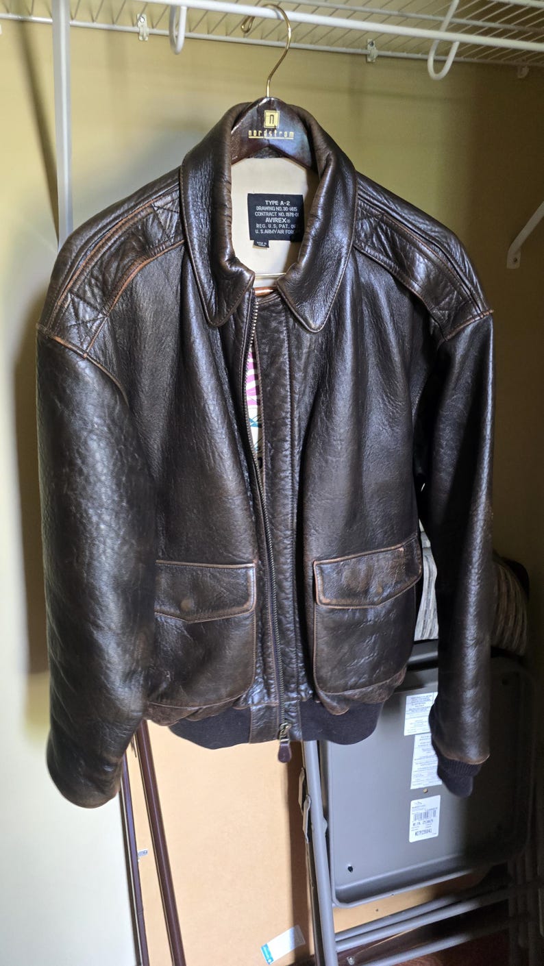 May include: A brown leather jacket with a zip-up front, two front pockets, and a ribbed hem and cuffs. The jacket is hanging on a hanger in a closet. The jacket has a collar and a label that reads "TYPE A-2".