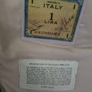 May include: Close-up of a vintage Allied Military Currency one lira note issued in Italy in 1943. The note is in a clear plastic sleeve, with a care instruction label below. The background is a light beige fabric.